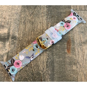 May include: A gray silicone Apple Watch band with a floral and hummingbird print. The band has a gold buckle.
