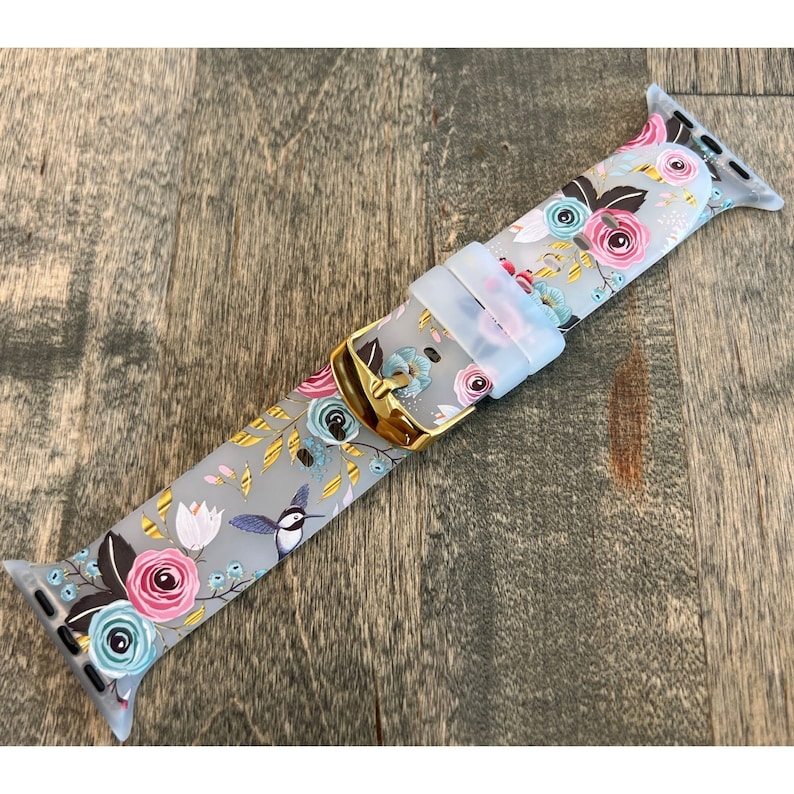 May include: A gray silicone Apple Watch band with a floral and hummingbird print. The band has a gold buckle.