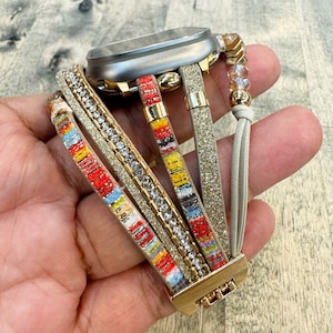 Może przedstawiać: Wielowarstwowy pasek do zegarka ze złotym zapięciem i tarczą. Pasek ma mieszankę faktur, w tym błyszczący złoty pasek, pasek z przezroczystymi kryształkami i kolorowe wzorzyste paski z czerwonymi, żółtymi i niebieskimi akcentami.