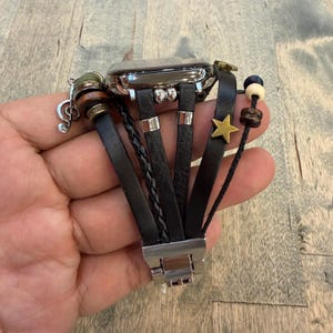 Puede incluir: Correa de cuero negro para Apple Watch con múltiples correas, un detalle trenzado y un cierre metálico. La correa presenta un dije de estrella dorada, cuentas de madera y un dije de nota musical.