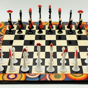 Pu&ograve; includere: Una scacchiera bianca e nera con un bordo circolare colorato. I pezzi degli scacchi sono bianchi e neri con accenti rossi e dorati. I pezzi sono disposti nelle loro posizioni di partenza.