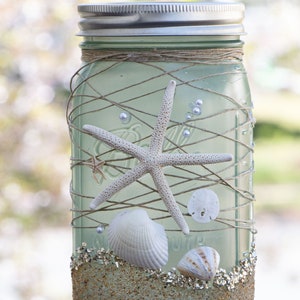 May include: A mason jar decorated with seashells, starfish, and twine. The jar is filled with sand and glitter, creating a beach-themed centerpiece.