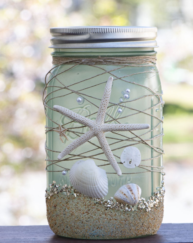 May include: A mason jar decorated with seashells, starfish, and twine. The jar is filled with sand and glitter, creating a beach-themed centerpiece.
