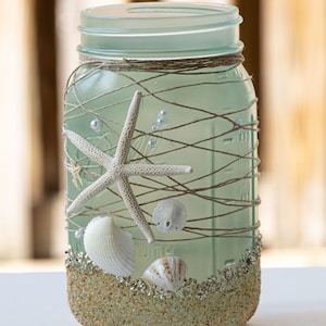 May include: A frosted glass mason jar decorated with twine, seashells, and starfish. The jar is filled with sand and glitter, creating a beach-themed design.