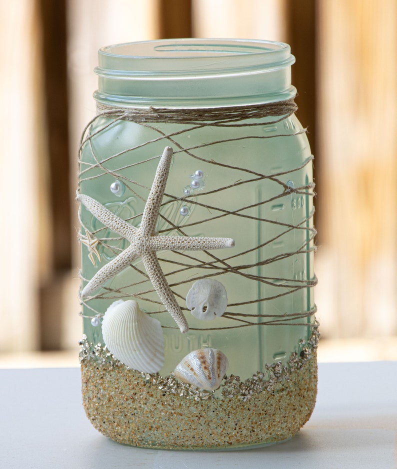 May include: A frosted glass mason jar decorated with twine, seashells, and starfish. The jar is filled with sand and glitter, creating a beach-themed design.
