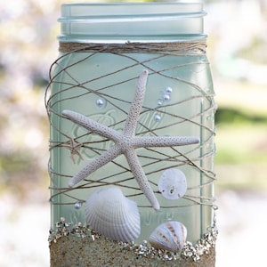 May include: A frosted glass mason jar decorated with twine, seashells, and a starfish. The jar is filled with sand and has a light blue tint.