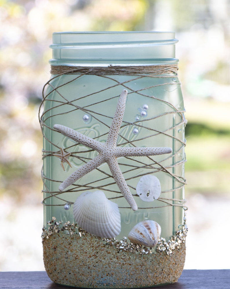 May include: A frosted glass mason jar decorated with twine, seashells, and a starfish. The jar is filled with sand and has a light blue tint.