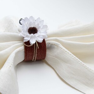 May include: A white linen napkin with a brown and white fabric napkin ring. The napkin ring is decorated with a white fabric flower.