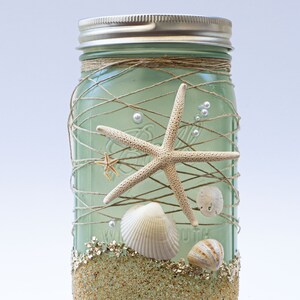 May include: A green glass mason jar decorated with twine, seashells, and starfish. The jar has a layer of gold glitter at the bottom, simulating sand.