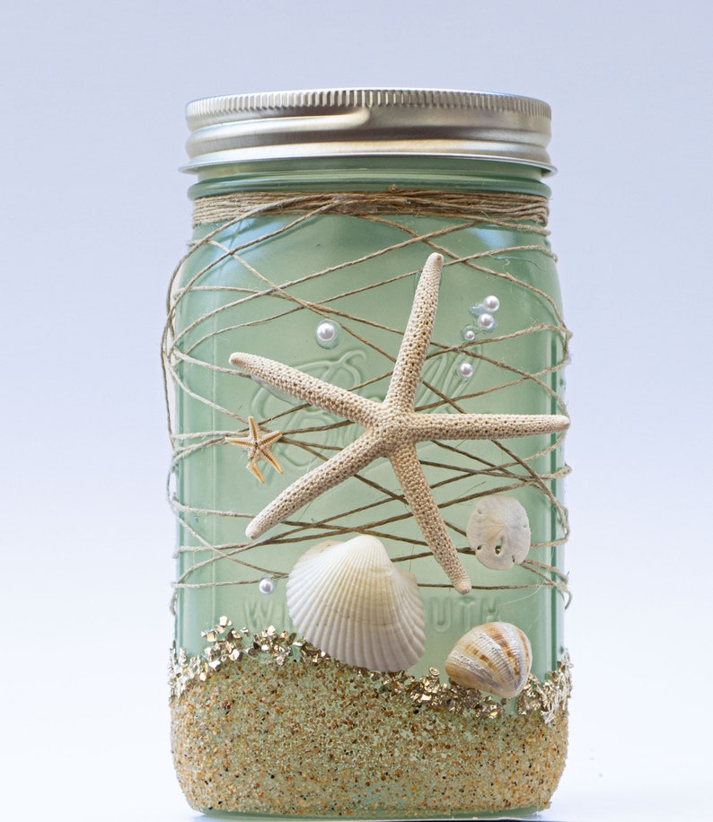 May include: A green glass mason jar decorated with twine, seashells, and starfish. The jar has a layer of gold glitter at the bottom, simulating sand.