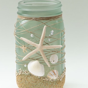 May include: A frosted green glass mason jar decorated with twine, seashells, and starfish. The jar is filled with sand and glitter, creating a beach-themed design.