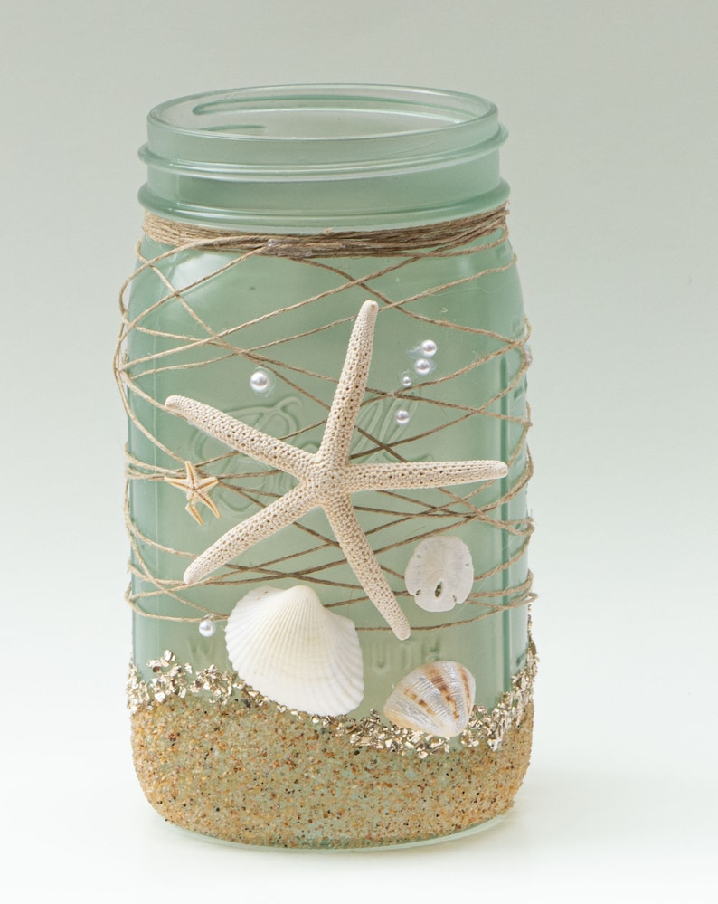 May include: A frosted green glass mason jar decorated with twine, seashells, and starfish. The jar is filled with sand and glitter, creating a beach-themed design.