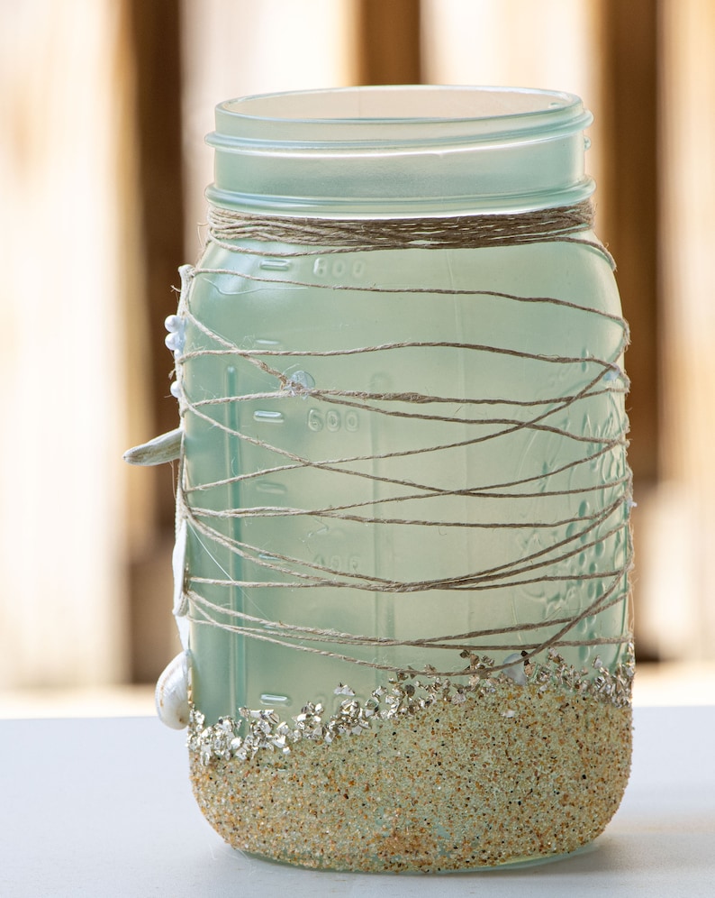 May include: A frosted glass jar decorated with twine and seashells. The bottom of the jar is covered in sand-colored glitter.