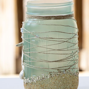May include: A frosted glass jar decorated with twine and seashells. The bottom of the jar is covered in sand-colored glitter.