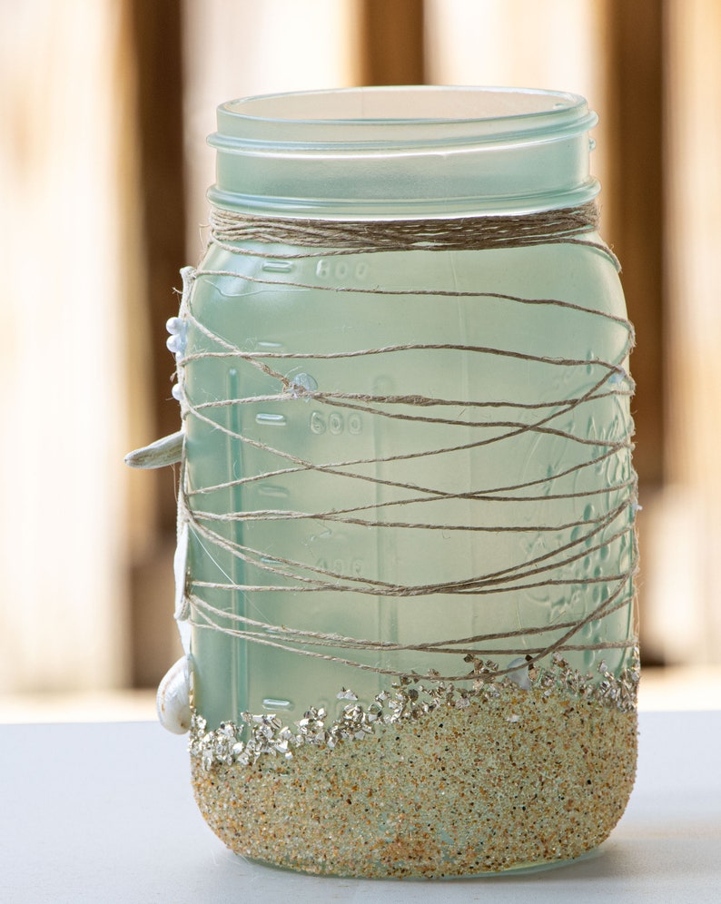 May include: A frosted glass jar decorated with twine and seashells. The bottom of the jar is covered in sand-colored glitter.
