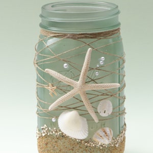 May include: A frosted glass mason jar decorated with twine, seashells, and starfish. The jar is filled with sand and has a light green tint.
