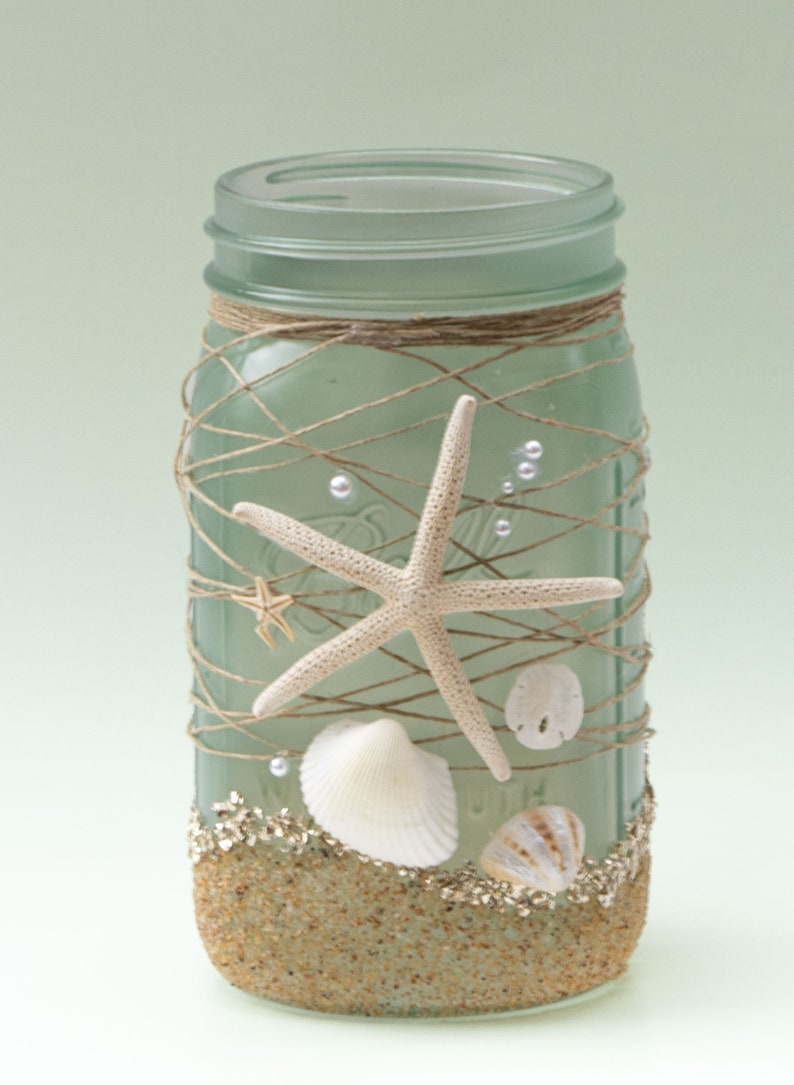 May include: A frosted glass mason jar decorated with twine, seashells, and starfish. The jar is filled with sand and has a light green tint.