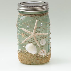 May include: A green glass mason jar decorated with twine, seashells, and starfish. The jar is filled with sand and has a silver lid.