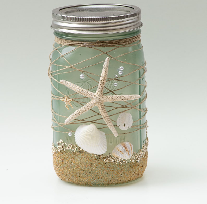 May include: A green glass mason jar decorated with twine, seashells, and starfish. The jar is filled with sand and has a silver lid.