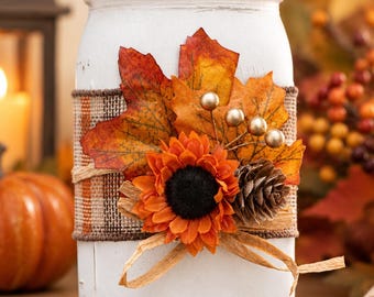 May include: A white painted glass mason jar with a silver metal lid, wrapped in a brown plaid burlap ribbon. The jar is decorated with orange and red maple leaves, a bright orange sunflower, a small pinecone, and gold-colored berry sprigs.