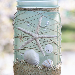 May include: A blue glass mason jar decorated with twine, seashells, and a starfish. The jar is filled with sand and glitter, creating a beach-themed design.