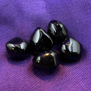 May include: Six black tumbled stones, likely obsidian, on a purple fabric background.