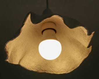 May include: A white, paper-like lampshade with a ruffled edge hangs from the ceiling. A light bulb illuminates the room from inside the shade.