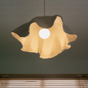 Op de afbeelding: Een witte lampenkap van papier met een golvende, organische vorm hangt aan het plafond. De lampenkap wordt verlicht door een enkele gloeilamp, die een warm licht geeft.