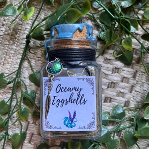 Peut inclure: Un petit bocal en verre avec un sceau de cire bleu et un couvercle en liège. Le bocal contient un petit charme iridescent et une étiquette qui indique "Occamy Eggshells".