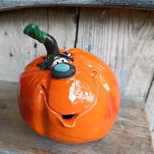 Könnte beinhalten: Eine glänzende orangefarbene Kürbisfigur aus Keramik mit einem breiten, lächelnden Mund, einer markanten blauen Nase und zwei weißen Augen mit schwarzen Pupillen. Ein dunkelgrüner, geschwungener Stiel ragt aus dem runden Kürbiskörper.