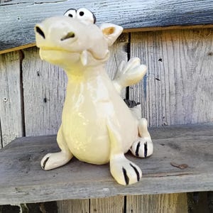 Könnte beinhalten: Eine skurrile, cremefarbene Keramik-Drachenfigur mit schwarzen Akzenten an Krallen und Augen. Der Drache hat kleine Flügel und posiert auf einem verwitterten Holzregal. Die Figur hat ein glänzendes Finish und einen verspielten Ausdruck.