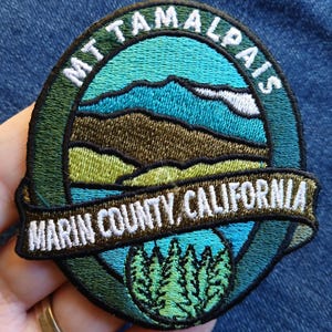 Pode incluir: Remendo bordado com um design de paisagem. Apresenta o texto "MT TAMALPAIS" e "MARIN COUNTY, CALIFORNIA". O remendo é circular com uma borda verde, e o design inclui montanhas, árvores e um céu azul.