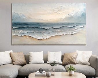 May include: Textured oil painting of ocean waves in shades of blue, white, and beige. The artwork depicts a seascape with a sandy beach and a cloudy sky. The painting is framed in a black border and hangs above a grey sofa with cushions.
