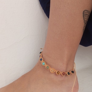 May include: A macrame ankle bracelet featuring a series of colorful gemstones. The bracelet is made with tan-colored cord and has gemstones in various colors, including blue, green, yellow, and red. The bracelet is worn on an ankle.