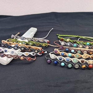 Puede incluir: Una colección de 10 pulseras de cuentas coloridas con una variedad de piedras, incluyendo rojo, azul, verde y marrón. Las pulseras están hechas con un cordón anudado y están dispuestas en una fila sobre una superficie blanca.