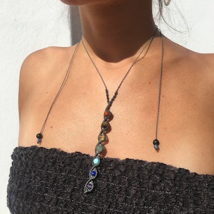 May include: A black macrame necklace with seven colorful gemstone beads. The beads are arranged in a vertical line and are spaced evenly apart. The necklace is worn by a person with a tan complexion.