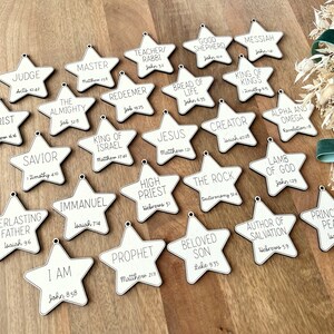May include: A set of 20 white wooden star ornaments with black text. Each star features a different title and scripture reference related to Jesus Christ. Titles include "Christ", "Judge", "Master", "Teacher/Rabbi", "Good Shepherd", "Messiah", "King of Kings", "The Almighty", "Savior", "Everlasting Father", "I Am", "Redeemer", "King of Israel", "Immanuel", "Bread of Life", "Jesus", "High Priest", "Prophet", "Creator", "The Rock", "Beloved Son", "Alpha and Omega", "Lamb of God", "Author of Salvation", "Prince of Peace".