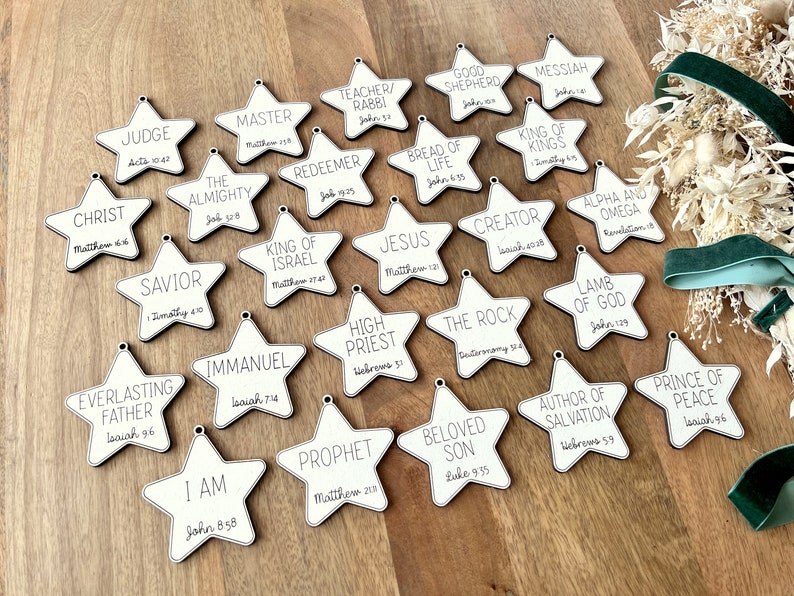 May include: A set of 20 white wooden star ornaments with black text. Each star features a different title and scripture reference related to Jesus Christ. Titles include "Christ", "Judge", "Master", "Teacher/Rabbi", "Good Shepherd", "Messiah", "King of Kings", "The Almighty", "Savior", "Everlasting Father", "I Am", "Redeemer", "King of Israel", "Immanuel", "Bread of Life", "Jesus", "High Priest", "Prophet", "Creator", "The Rock", "Beloved Son", "Alpha and Omega", "Lamb of God", "Author of Salvation", "Prince of Peace".