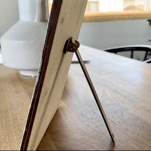 Op de afbeelding: Een houten fotolijst met een metalen standaard. De lijst is lichtbruin en heeft een witte achtergrond. De standaard is zilverkleurig en klapt uit om de lijst rechtop te zetten.