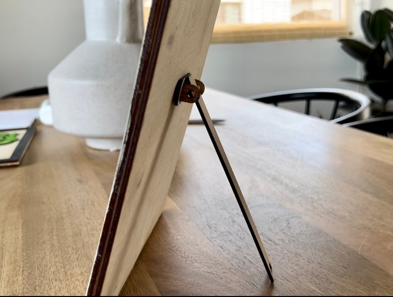 Op de afbeelding: Een houten fotolijst met een metalen standaard. De lijst is lichtbruin en heeft een witte achtergrond. De standaard is zilverkleurig en klapt uit om de lijst rechtop te zetten.