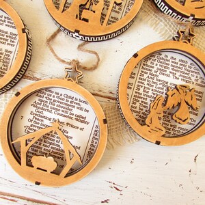 May include: Wooden Christmas ornaments with a circular design. Each ornament features a cutout nativity scene or angel, with a star-shaped hanger. The ornaments have a light brown color and contain text from a book.