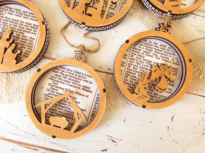 May include: Wooden Christmas ornaments with a circular design. Each ornament features a cutout nativity scene or angel, with a star-shaped hanger. The ornaments have a light brown color and contain text from a book.