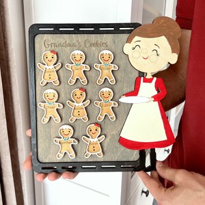K&ouml;nnte beinhalten: Ein Holzschild mit dem Text "Grandma's Cookies" und einem Ausschnitt einer Frau, die einen Teller h&auml;lt. Es gibt acht Lebkuchenm&auml;nner mit darauf geschriebenen Namen.