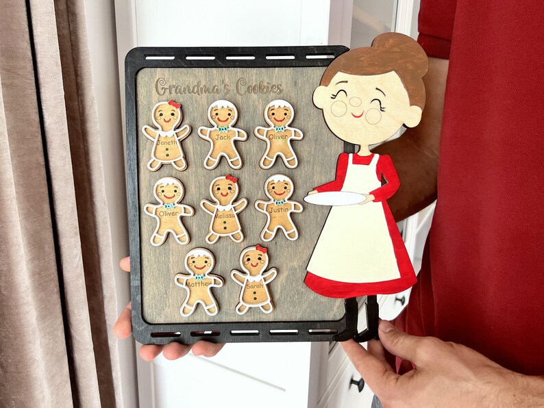 K&ouml;nnte beinhalten: Ein Holzschild mit dem Text "Grandma's Cookies" und einem Ausschnitt einer Frau, die einen Teller h&auml;lt. Es gibt acht Lebkuchenm&auml;nner mit darauf geschriebenen Namen.