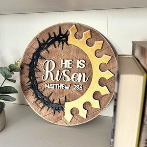 May include: A round wooden sign with a brown background and a chevron pattern. The sign features a black crown of thorns, a gold crown, and the text "He is Risen" with the scripture reference "Matthew 28:6".
