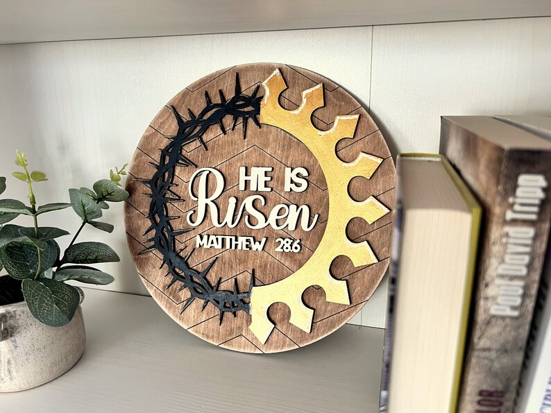 May include: A round wooden sign with a brown background and a chevron pattern. The sign features a black crown of thorns, a gold crown, and the text "He is Risen" with the scripture reference "Matthew 28:6".