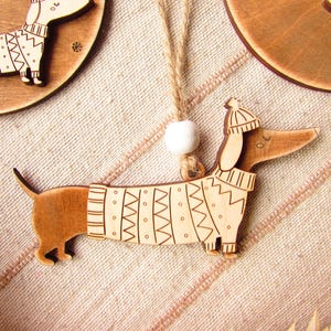 May include: Wooden dachshund ornament wearing a sweater and hat. The dog is light brown with etched details, and the sweater has a geometric pattern. A white bead and twine are attached for hanging. Other similar ornaments are in the background.