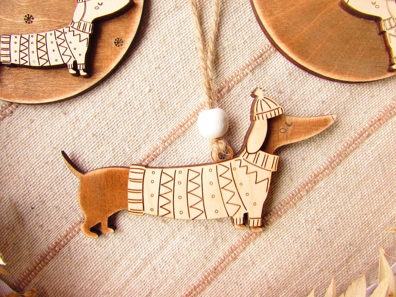 May include: Wooden dachshund ornament wearing a sweater and hat. The dog is light brown with etched details, and the sweater has a geometric pattern. A white bead and twine are attached for hanging. Other similar ornaments are in the background.