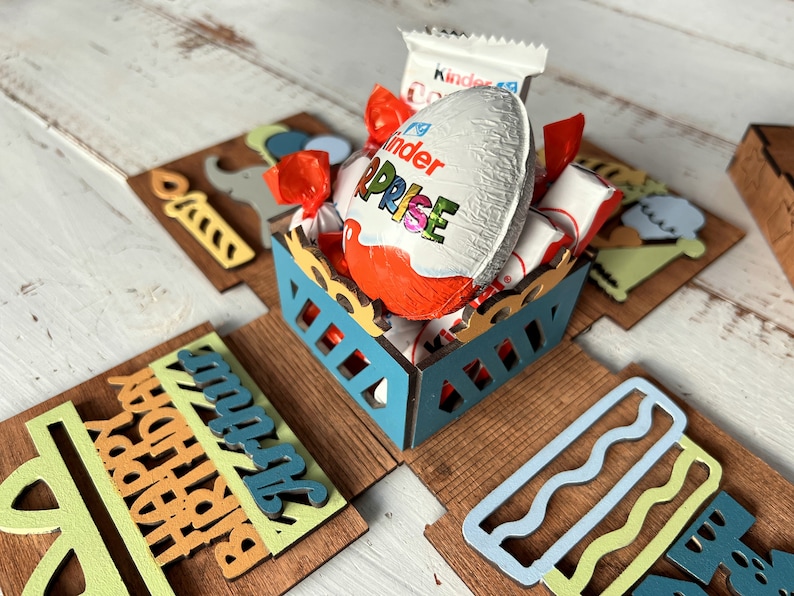 May include: A wooden box with a blue and yellow design. The box is open and filled with chocolate candies, including a Kinder Surprise egg. The box has a Happy Birthday message on the front.