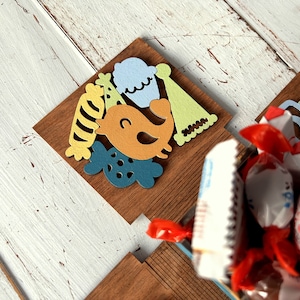 May include: A wooden box with a colorful birthday theme. The box is decorated with a bird, a cupcake, a party hat, and a striped candy. The box is filled with wrapped candies.
